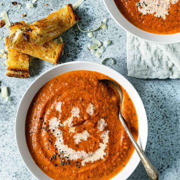 A bowl of tomato soup on a light grey background, with grilled cheese sticks on the side