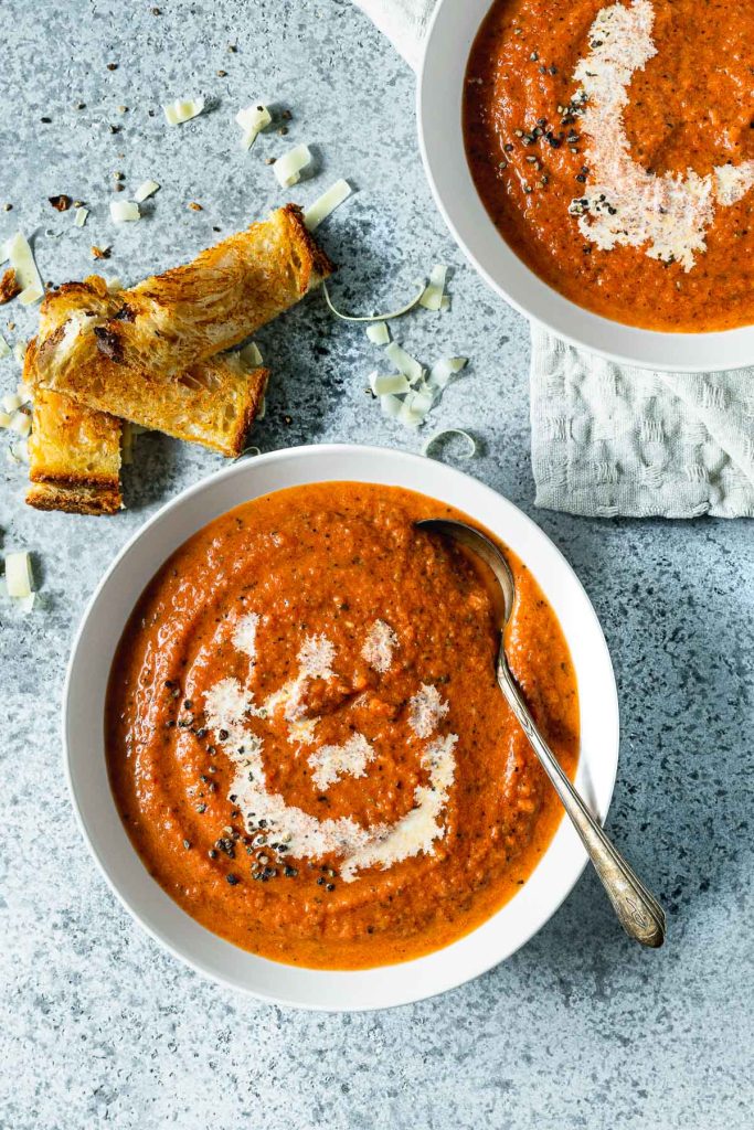 A bowl of tomato soup on a light grey background, with grilled cheese sticks on the side
