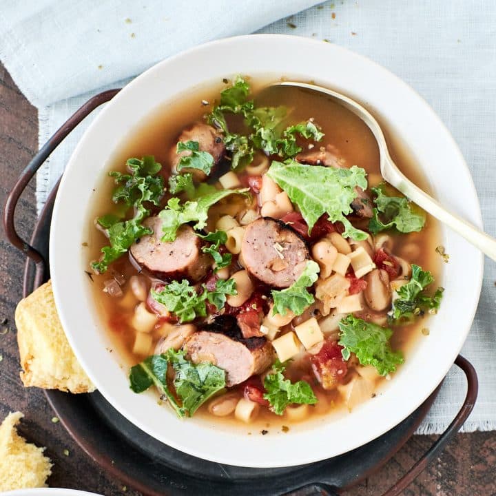 A bowl of tomato based soup with white beans, sausage and kale