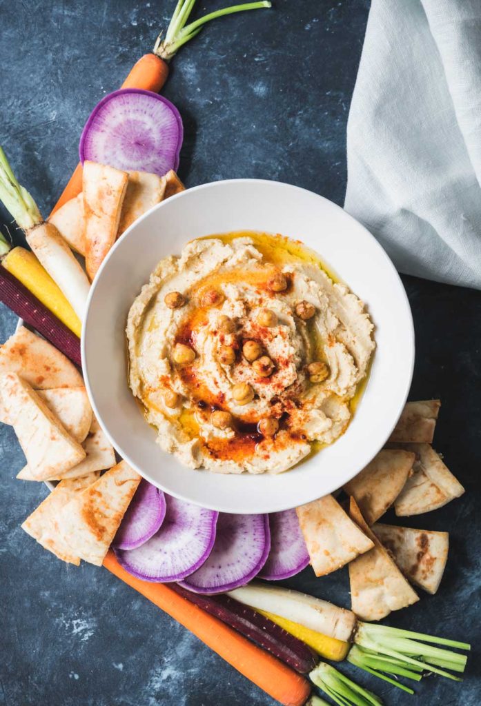 Hummus in a cream bowl with pita chips and bright veggies on the side