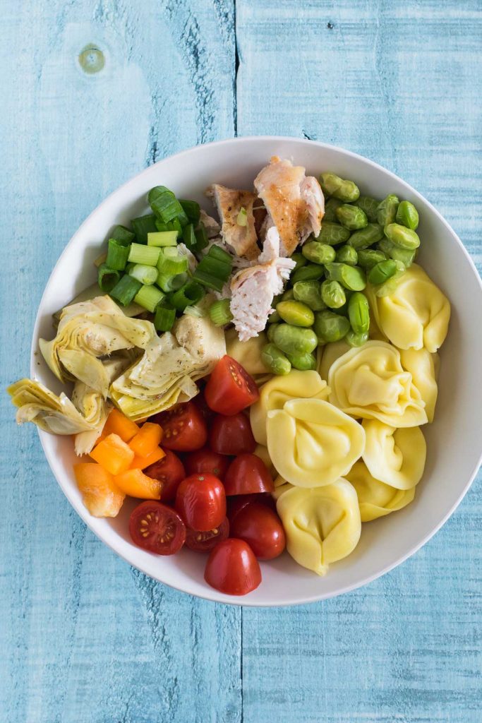 White bowl with cooked tortellini, chopped tomatoes, edamame and artichoke hearts