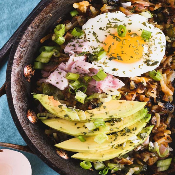 Skillet filled with hash browns, ham, sliced avocado and a poached egg