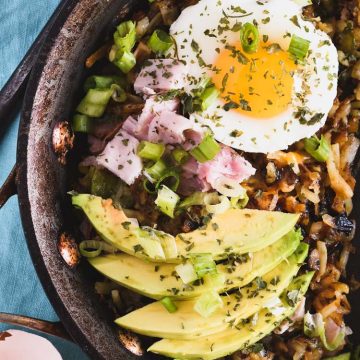 Skillet filled with hash browns that are topped with ham, a poached egg and avocado slices
