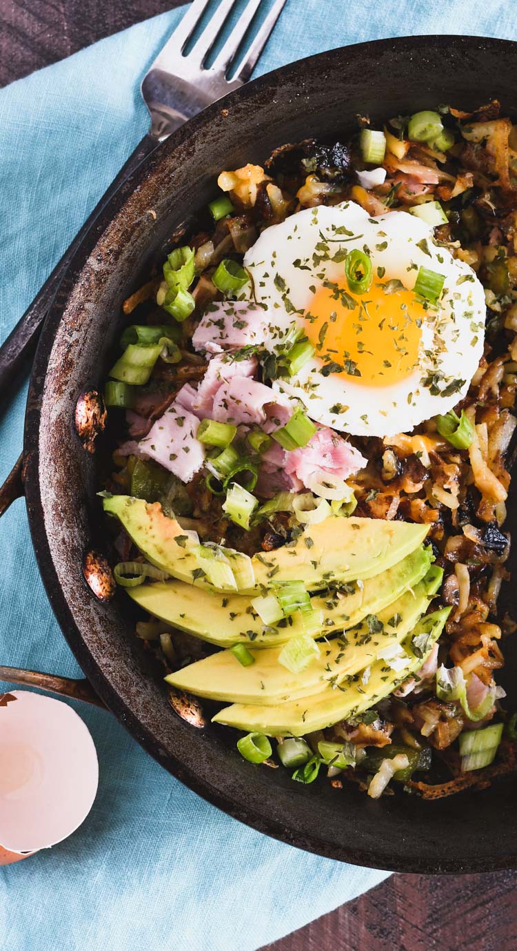 A pan filled with hash brown potatoes topped with sliced ham, green onions, avocado and a poached egg