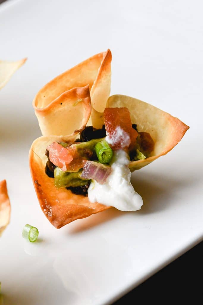 Close up of a wonton cup on a white platter filled with black beans, guacamole and pico de gallo