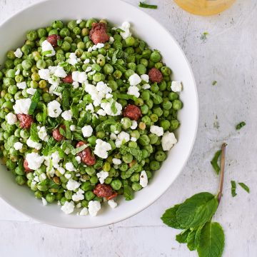 Overhead shot of english peas in a white bowl topped with goat cheese and candied hazelnute=s