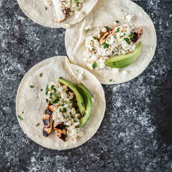 Three grilled chicken tacos topped with elotes and avocados