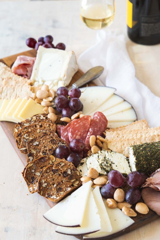 Cheese, meat and crackers on a dark brown board with a glass of wine in the background