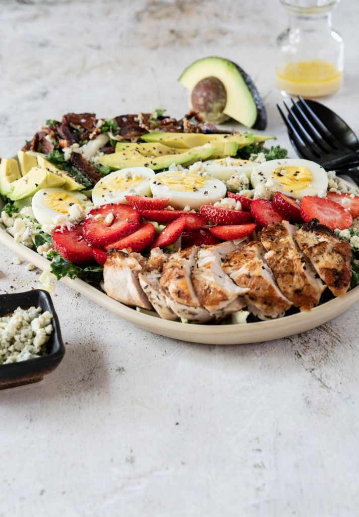 Cobb salad topped with chicken, avocado, bacon and sliced strawberries on a cream platter