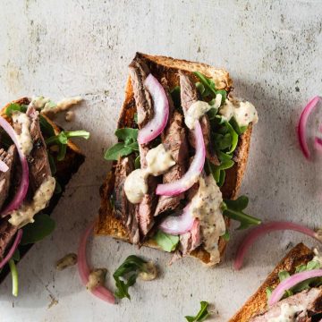 Close up of an open faced steak sandwich on crusty bread topped with bright pink pickled onions