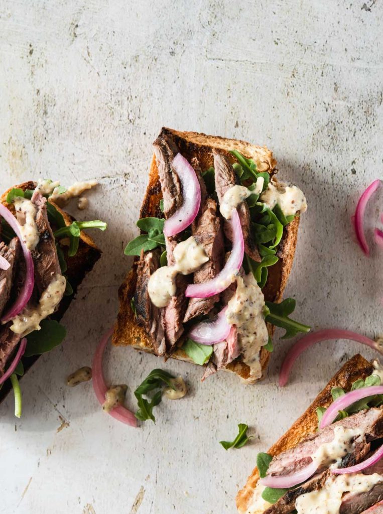 Close up of an open faced steak sandwich on crusty bread topped with bright pink pickled onions