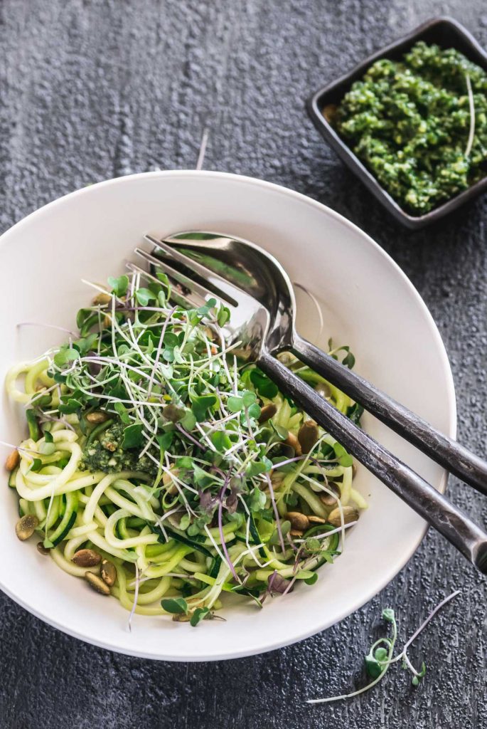 Overhead shot of zucchini noodles in a white bowl topped with pesto and micro greens