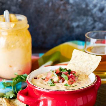 Queso dip in a red bowl with tortilla chips scattered in front