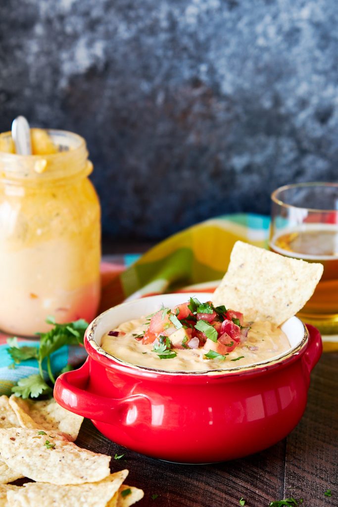 Queso dip in a red bowl with tortilla chips scattered in front