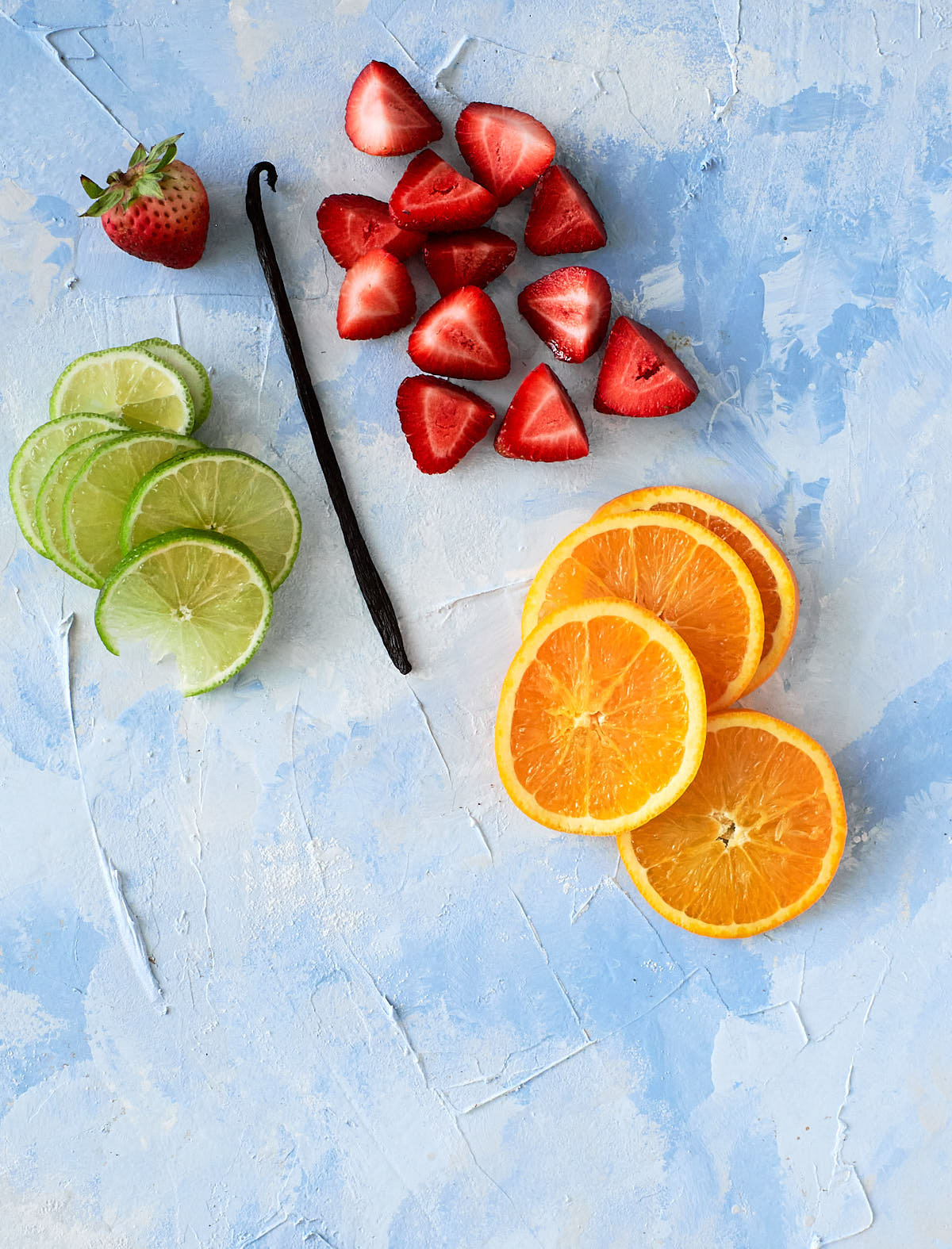 Slices of orange, lime and strawberry plus a vanilla bean, on a light blue background
