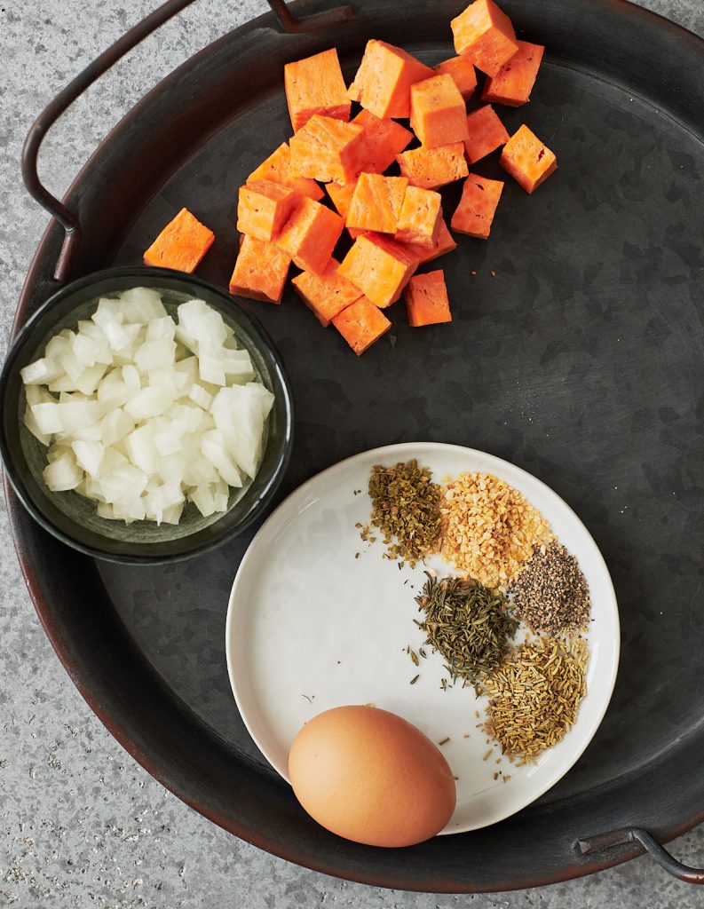 Cubed sweet potatoes, chopped onions, a brown egg and spices on a black metal tray