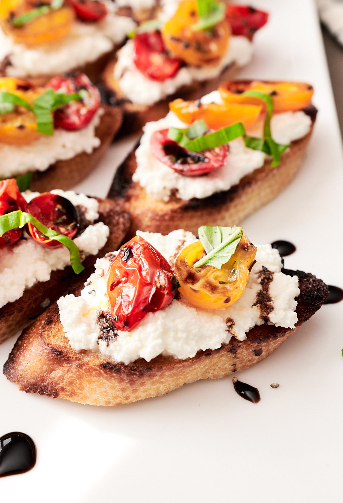 Close up shot of a slice of grilled bread topped with ricotta, roasted tomatoes and balsamic glaze