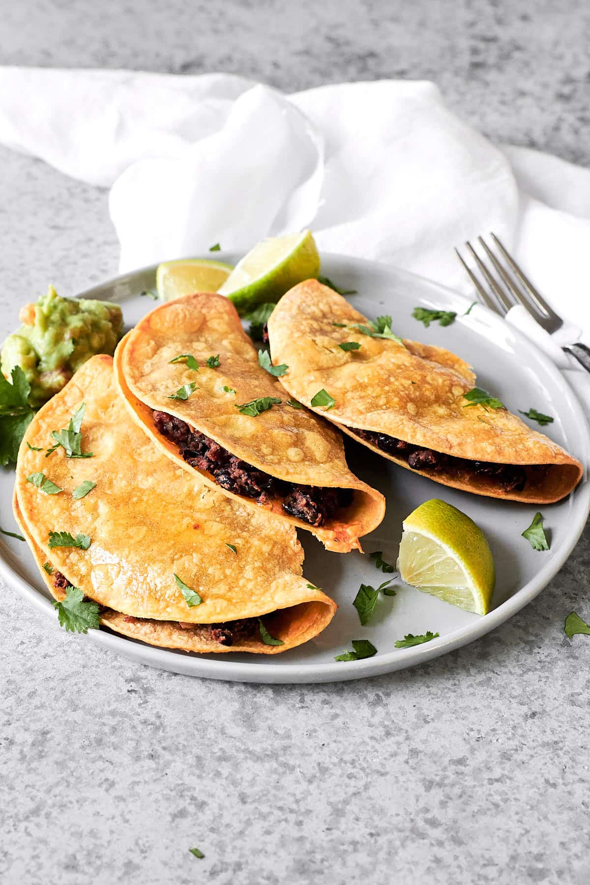 Three corn tortillas stuffed with black beans with a side of guacamole and some lime wedges