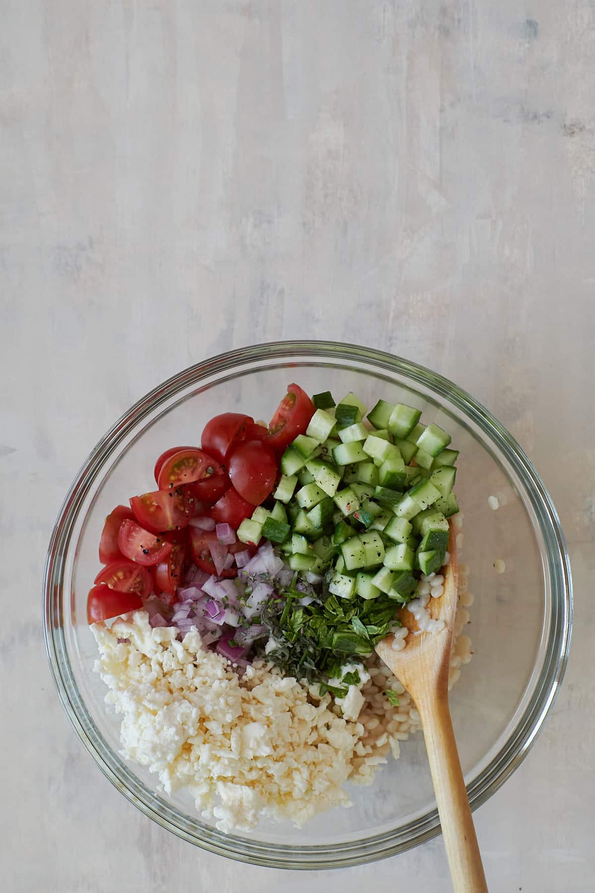 Glass bowl filled with pearled couscous, grape tomatoes, red onion, feta and diced cucumber