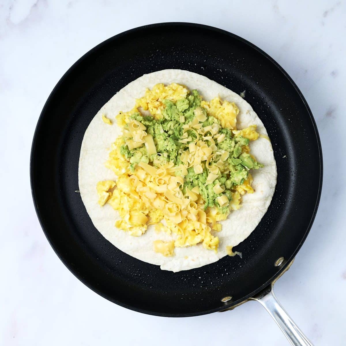 Tortilla in a pan topped with scrambled eggs, smashed avocado and shredded cheese