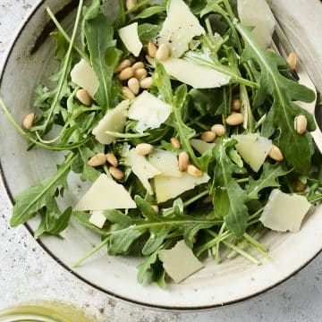 Small salad bowl filled with arugula topped with pine nuts and shaved parmesan cheese
