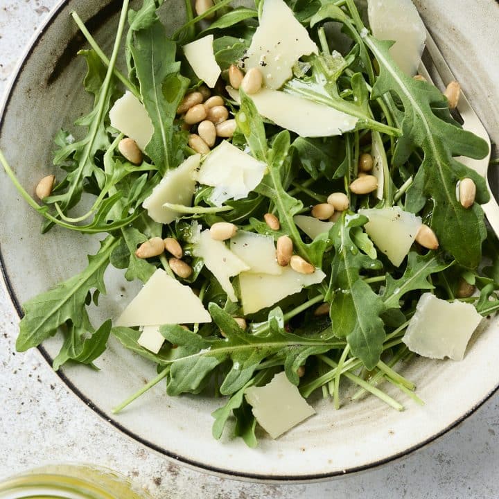 Small salad bowl filled with arugula topped with pine nuts and shaved parmesan cheese