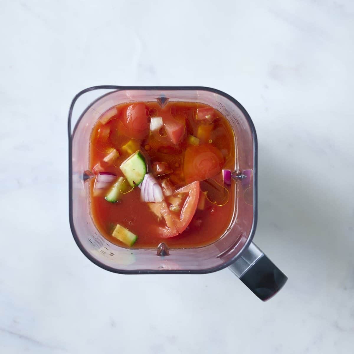 Top-down view of a blender containing chopped tomatoes, cucumbers, red onions and tomato juice for tomato gazpacho, ready to be blended, on a white marble surface.